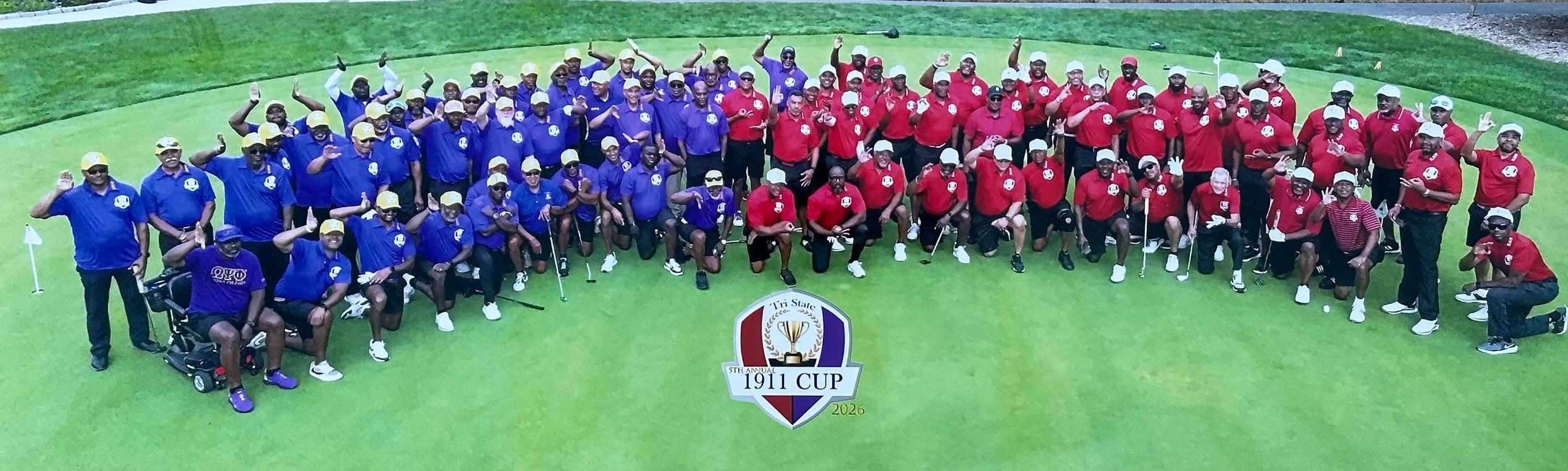 Aerial photo of the 5th annual Tri State 1911 Cup field, 2026: Omega Psi Phi golfers in purple and Kappa Alpha Psi golfers in red gathered on the putting green around the Cup logo